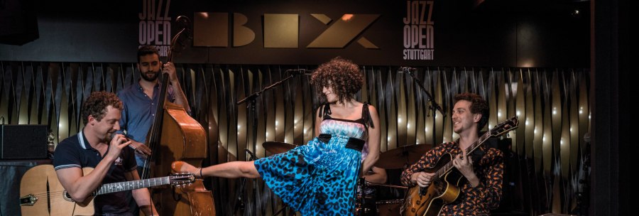 A band plays in the BIX jazz club. A guitarist, a double bass player and a singer in a blue dress can be seen. "Jazz Open Stuttgart" in the background., © Peter Steinheisser