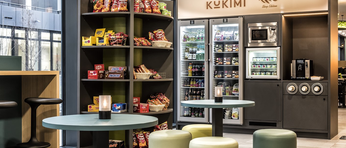 Moderne Lobby mit Snackregal, Kühlschrank und Sitzbereich. Der Raum ist hell und einladend gestaltet, mit runden Tischen und Hockern., © ibis Styles Stuttgart Vaihingen