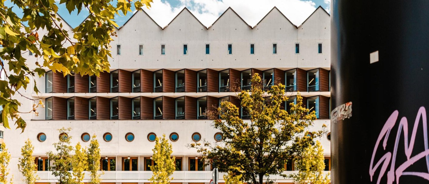 Moderne Bibliothek mit markanter geometrischer Fassade, umgeben von Bäumen. Ein schwarzer Pfeiler mit Graffiti ist rechts im Bild sichtbar., © Stuttgart Marketing GmbH, Sarah Schmid Moderne Bibliothek mit markanter geometrischer Fassade, umgeben von Bäumen. Ein schwarzer Pfeiler mit Graffiti ist rechts im Bild sichtbar., © Stuttgart Marketing GmbH, Sarah Schmid
