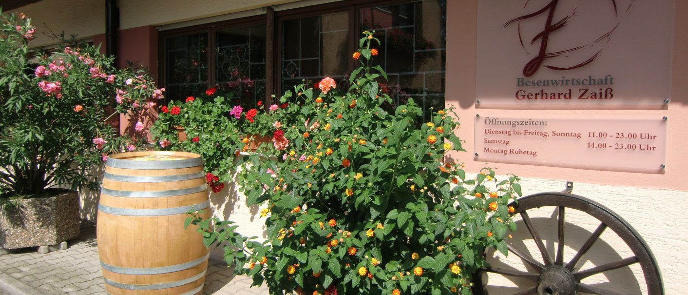Entrance to the Besenwirtschaft Gerhard Zaiß with flowering plants, a wine barrel and a sign with opening hours., © Simone Mack Entrance to the Besenwirtschaft Gerhard Zaiß with flowering plants, a wine barrel and a sign with opening hours., © Simone Mack