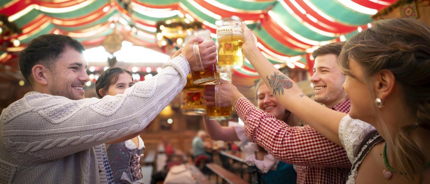 People in traditional dress clink glasses with mugs of beer in a festively decorated tent. Cheerful atmosphere at a folk festival., © Stuttgart-Marketing GmbH, Sarah Schmid People in traditional dress clink glasses with mugs of beer in a festively decorated tent. Cheerful atmosphere at a folk festival., © Stuttgart-Marketing GmbH, Sarah Schmid