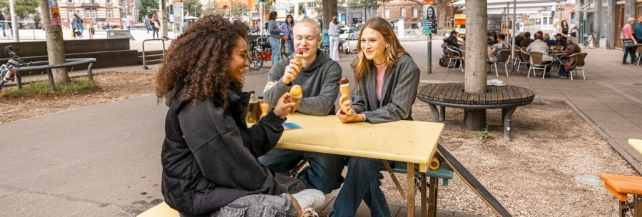 Three people are sitting at a yellow table outside eating ice cream. Trees, bicycles and a café can be seen in the background., © SMG Stuttgart Marketing GmbH - Sarah Schmid