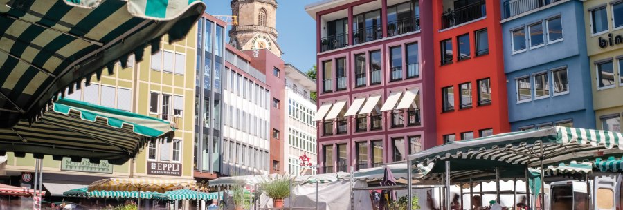 Bunte Geb&auml;ude und Marktst&auml;nde auf einem Marktplatz, mit einem Kirchturm im Hintergrund unter blauem Himmel., &copy; SMG, trickytine