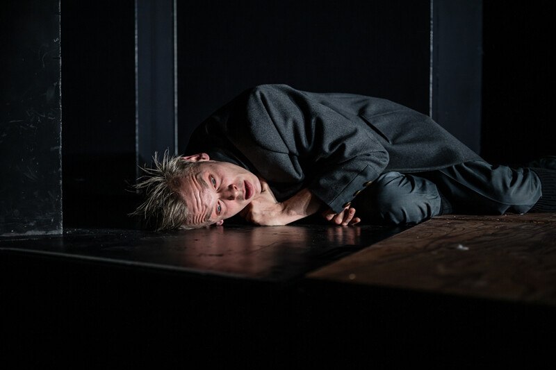 Ein Schauspieler in einem dunklen Raum liegt auf dem Boden und schaut nachdenklich in die Kamera. Die Szene wirkt dramatisch und intensiv., © Württembergische Staatstheater Stuttgart Ein Schauspieler in einem dunklen Raum liegt auf dem Boden und schaut nachdenklich in die Kamera. Die Szene wirkt dramatisch und intensiv., © Württembergische Staatstheater Stuttgart