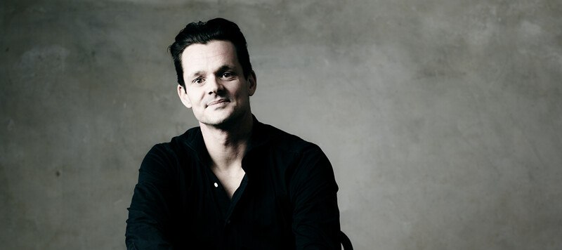 A man in a black shirt sits in front of a gray background and smiles slightly into the camera., &copy; W&uuml;rttembergische Staatstheater Stuttgart