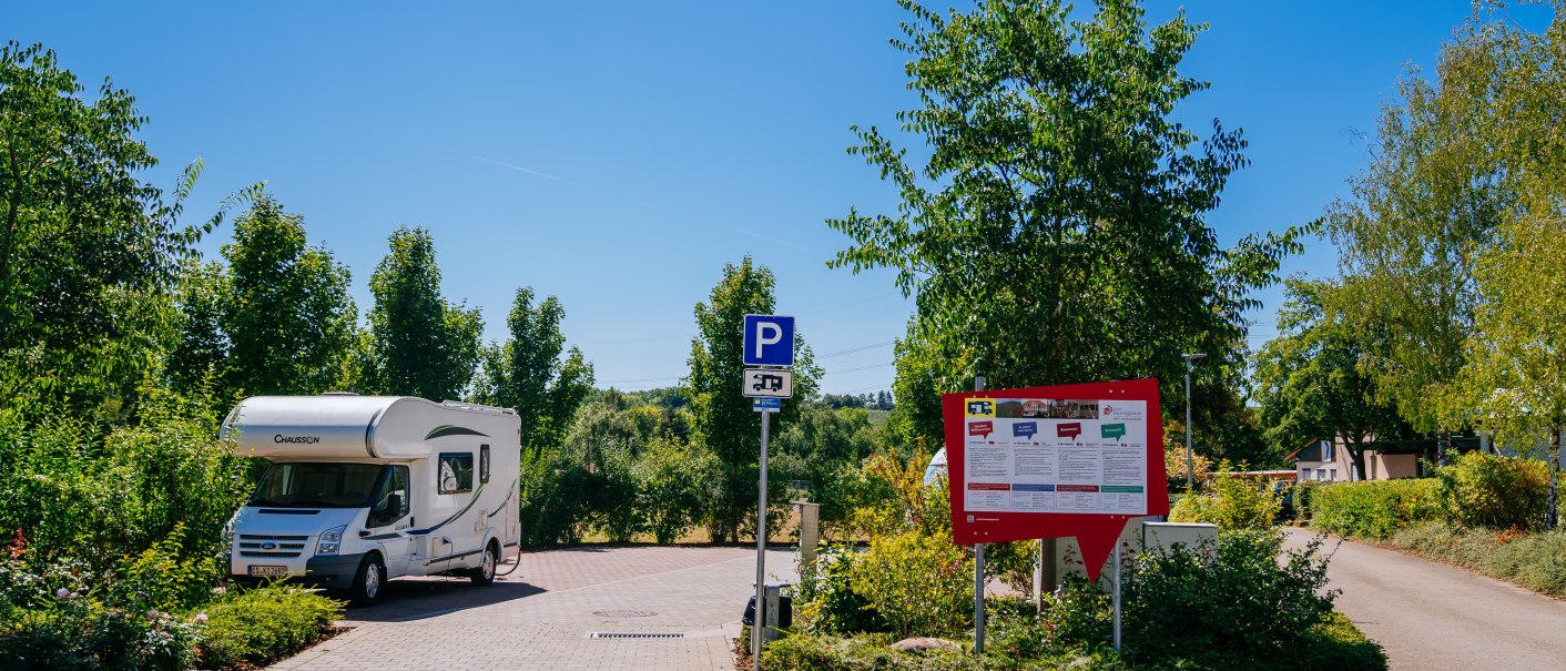 Wohnmobilstellplatz in Bönnigheim mit einem geparkten Wohnmobil, umgeben von Bäumen und einem Informationsschild bei klarem Himmel., © Stuttgart-Marketing GmbH, Thomas Niedermüller