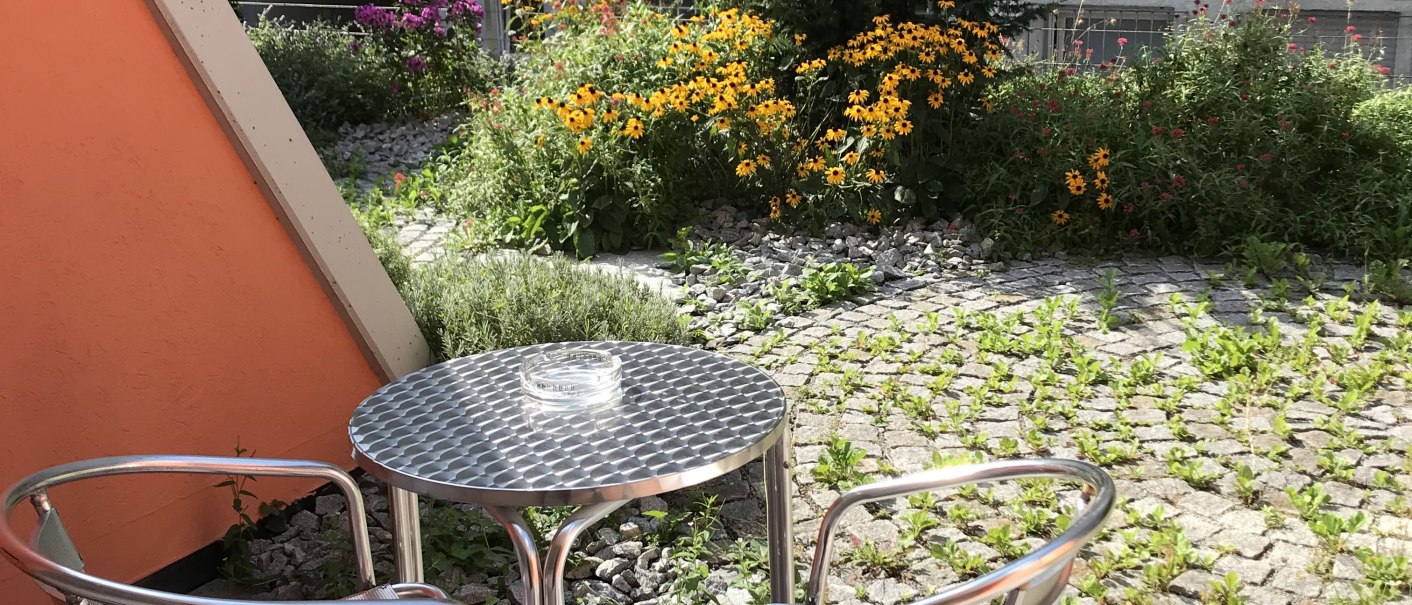 Kleine Terrasse mit zwei Metallstühlen und einem Tisch. Im Hintergrund ein Garten mit Blumen und ein graues Gebäude., © Abalon Hotel Ideal Kleine Terrasse mit zwei Metallstühlen und einem Tisch. Im Hintergrund ein Garten mit Blumen und ein graues Gebäude., © Abalon Hotel Ideal