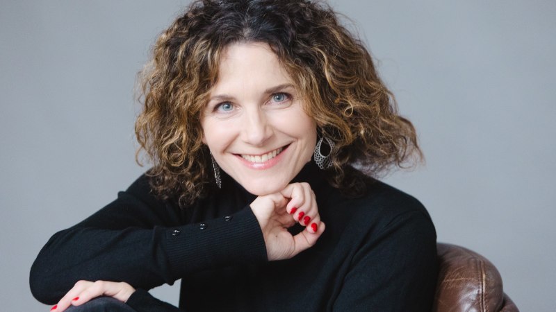 Eine Frau mit lockigem Haar und Ohrringen l&auml;chelt in die Kamera. Sie tr&auml;gt einen schwarzen Pullover und sitzt auf einem braunen Ledersessel., &copy; Uwe Hauth