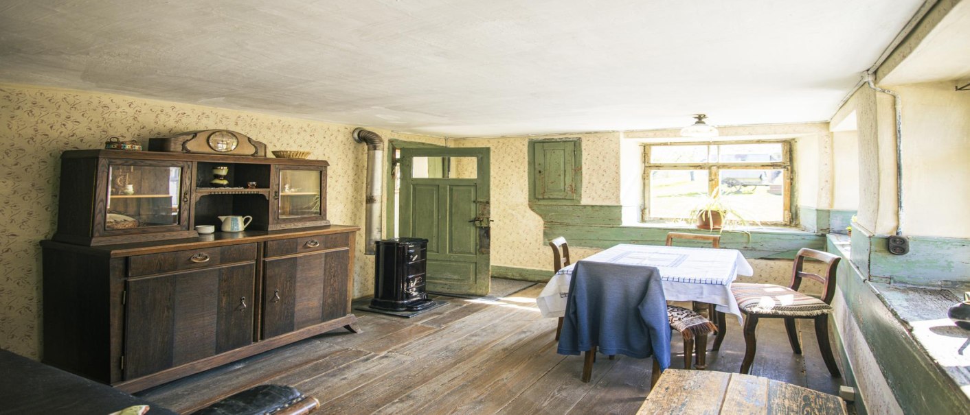 Ein rustikales Wohnzimmer mit Holzboden, antikem Schrank, Tisch mit Decke, Stühlen und einem Ofen. Sonnenlicht fällt durch das Fenster., © SMG, Sarah Schmid