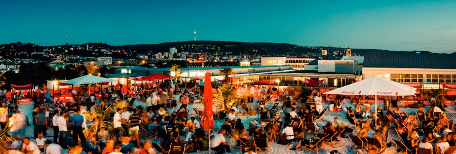 Large crowd on a roof terrace at dusk, surrounded by parasols and deckchairs, with a view of an illuminated city., &copy; Sky Beach