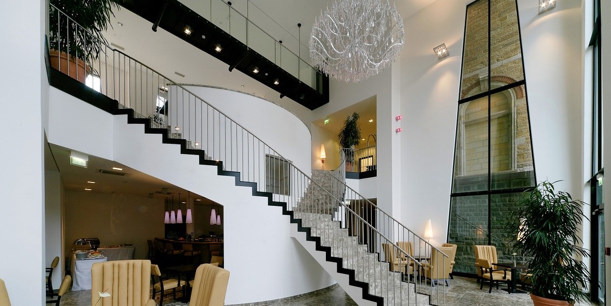 Elegant lobby with curved staircase, large chandelier and cozy seating area. High windows and plants create an inviting atmosphere., © Arcotel Hotels Elegant lobby with curved staircase, large chandelier and cozy seating area. High windows and plants create an inviting atmosphere., © Arcotel Hotels