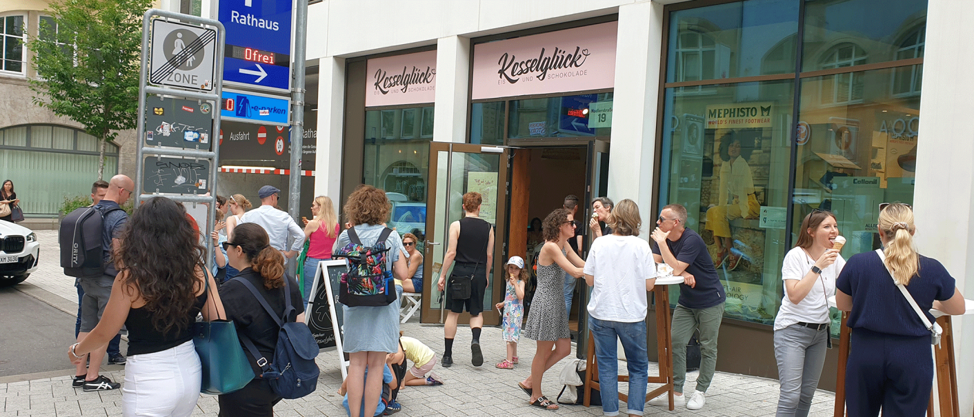 Menschen genießen Eis vor dem Eiscafé Kesselglück in einer belebten Stadtstraße. Einige stehen, andere sitzen an Tischen., © kesselglueck.de