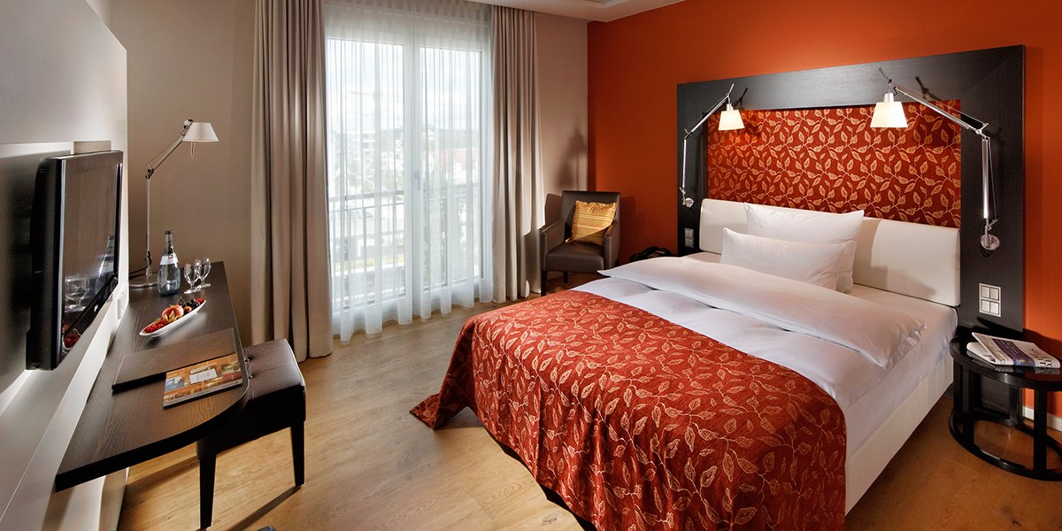 Elegantes Hotelzimmer mit roter Akzentwand, Doppelbett, Schreibtisch, Fernseher und gro&szlig;em Fenster mit Vorh&auml;ngen., &copy; Parkhotel Stuttgart Messe-Airport