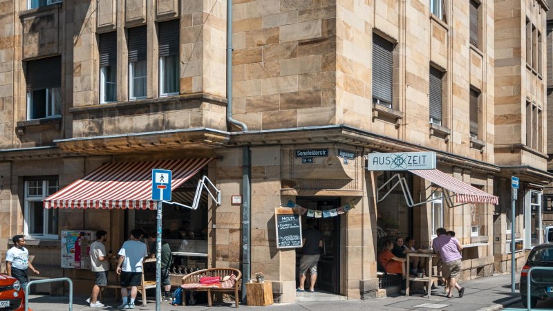 Cafébar Auszeit an einer Straßenecke mit roter Markise. Menschen sitzen draußen, Straßenschilder und Autos sind sichtbar., © SMG, Sarah Schmid Cafébar Auszeit an einer Straßenecke mit roter Markise. Menschen sitzen draußen, Straßenschilder und Autos sind sichtbar., © SMG, Sarah Schmid