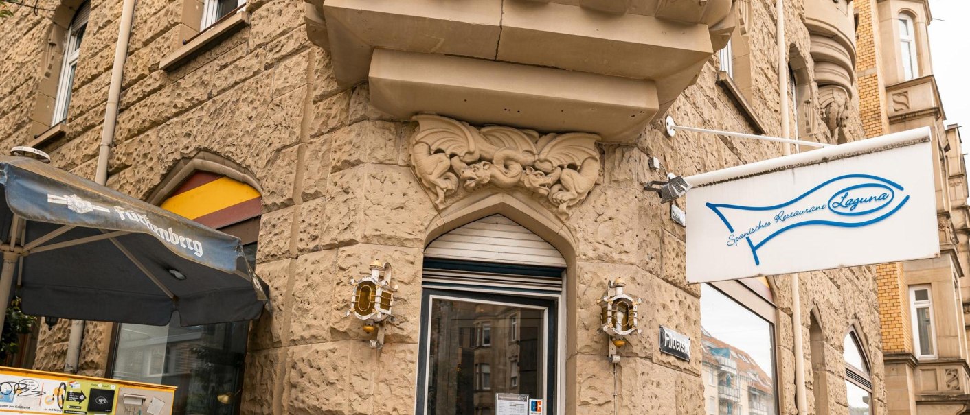 Eingang eines spanischen Restaurants namens Laguna mit Fisch-Logo. Sandsteinfassade mit dekorativen Elementen und einem Sonnenschirm., © SMG, Sarah Schmid Eingang eines spanischen Restaurants namens Laguna mit Fisch-Logo. Sandsteinfassade mit dekorativen Elementen und einem Sonnenschirm., © SMG, Sarah Schmid