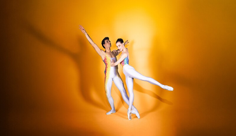 Zwei Ballettt&auml;nzer in bunten Kost&uuml;men posieren anmutig vor einem leuchtend gelben Hintergrund., &copy; W&uuml;rttembergische Staatstheater Stuttgart