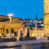 Evening atmosphere at Kleiner Schlossplatz with palm trees, people and modern architecture. Illuminated buildings and a statue can be seen in the background., &copy; SMG, Werner Dieterich