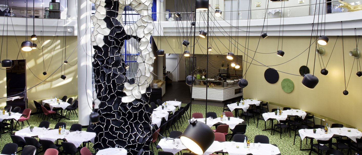 Ein modernes Restaurant mit k&uuml;nstlerischer Dekoration, h&auml;ngenden Lampen und gedeckten Tischen auf gr&uuml;nem Teppichboden., &copy; Holiday Inn