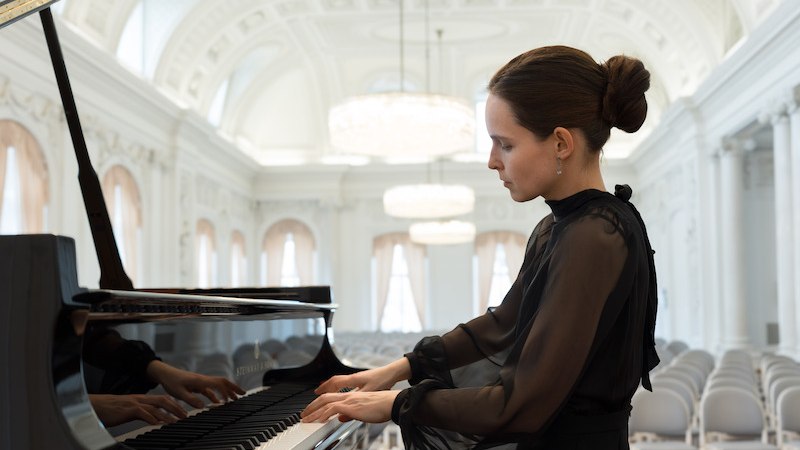 Eine Frau in schwarzer Kleidung spielt Klavier in einem eleganten, leeren Konzertsaal mit großen Kronleuchtern., © Amati Kammermusikschule Stuttgart e.V.