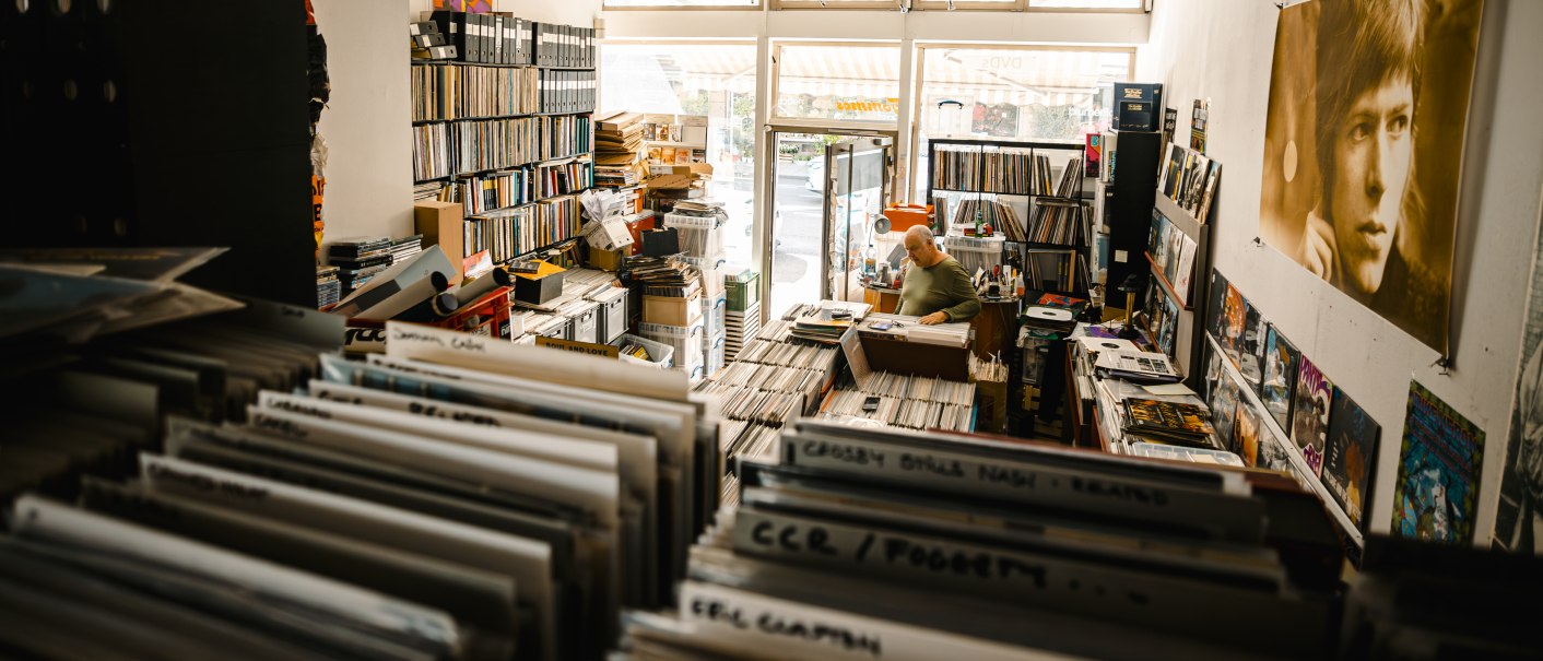 Ein gemütlicher Plattenladen voller Schallplatten. Ein Mann sitzt an einem Schreibtisch, umgeben von Regalen und Postern an den Wänden., © SMG Stuttgart Marketing GmbH - Sarah Schmid