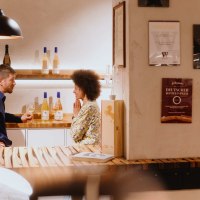 Zwei Personen unterhalten sich in einer Vinothek. Weinflaschen stehen auf Regalen, Urkunden an der Wand. Gemütliche Atmosphäre mit Holztischen., © SMG, Alwin Maigler Zwei Personen unterhalten sich in einer Vinothek. Weinflaschen stehen auf Regalen, Urkunden an der Wand. Gemütliche Atmosphäre mit Holztischen., © SMG, Alwin Maigler