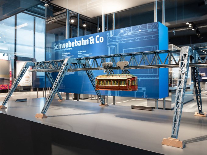A model of a suspension railroad in the Märklineum, displayed on a table. A blue information stand can be seen in the background., © Märklin Spielwaren Vertriebs-GmbH A model of a suspension railroad in the Märklineum, displayed on a table. A blue information stand can be seen in the background., © Märklin Spielwaren Vertriebs-GmbH
