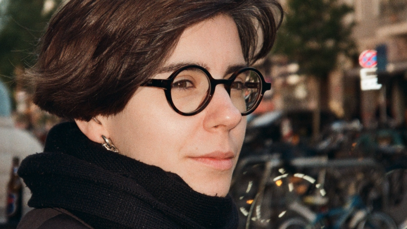 Eine Person mit Brille und schwarzem Schal steht in einer städtischen Umgebung. Im Hintergrund sind Fahrräder und Gebäude zu sehen., © privat Eine Person mit Brille und schwarzem Schal steht in einer städtischen Umgebung. Im Hintergrund sind Fahrräder und Gebäude zu sehen., © privat