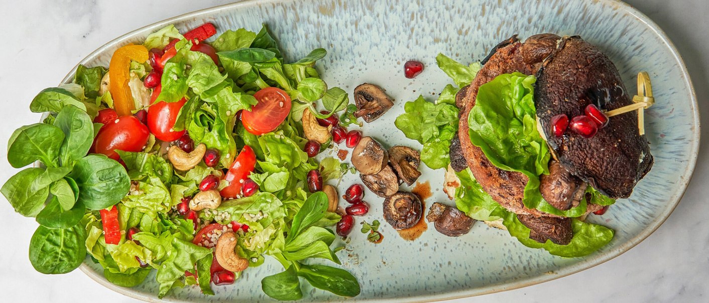 A plate with a salad of green leafy vegetables, tomatoes, peppers, cashew nuts and pomegranate seeds. Next to it, a burger with mushrooms., © SMG A plate with a salad of green leafy vegetables, tomatoes, peppers, cashew nuts and pomegranate seeds. Next to it, a burger with mushrooms., © SMG