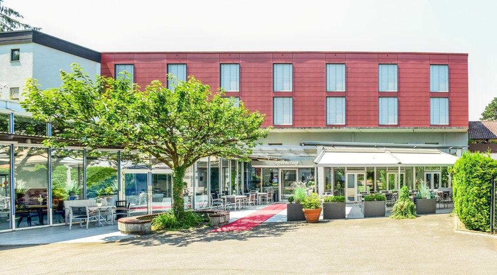 Modern building with red façade and glass front, surrounded by plants and terrace with seating., © Hotel Fuchsen Modern building with red façade and glass front, surrounded by plants and terrace with seating., © Hotel Fuchsen