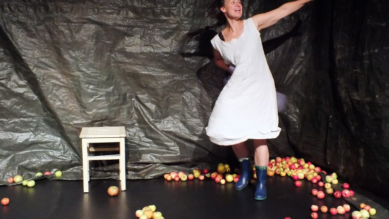 Eine Frau in einem wei&szlig;en Kleid und blauen Gummistiefeln tanzt auf einer B&uuml;hne, die mit &Auml;pfeln bedeckt ist. Ein Hocker steht im Hintergrund., &copy; Dieter Nelle