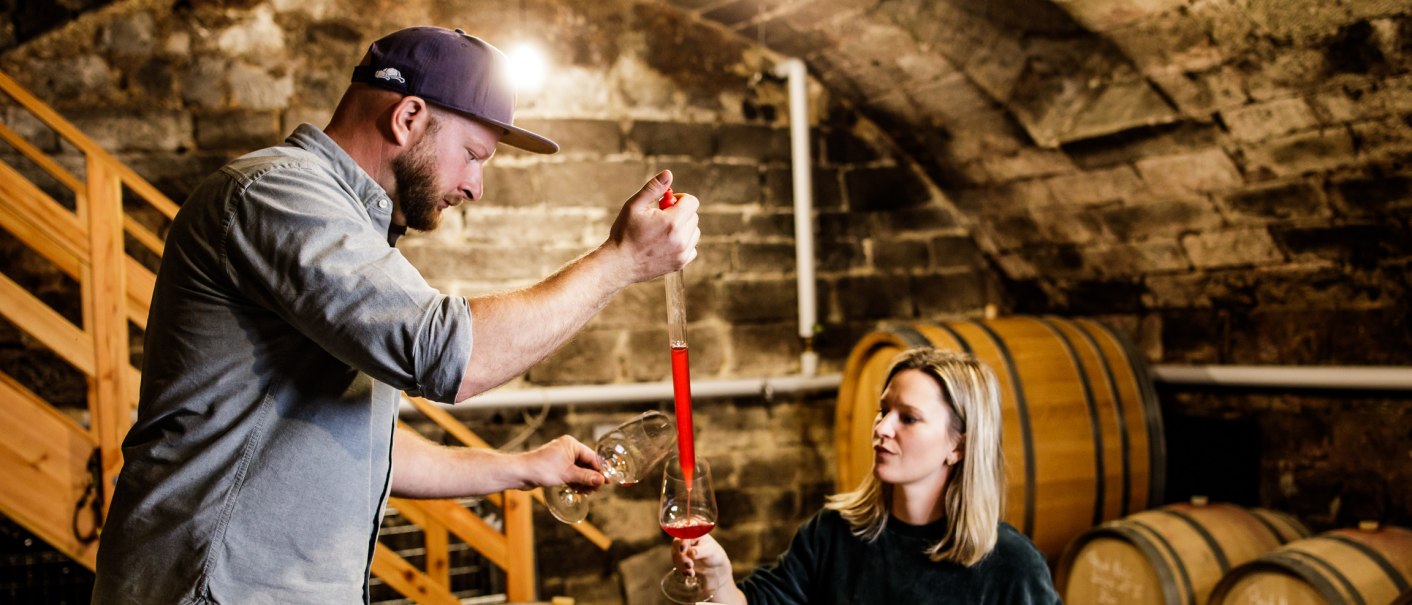 Zwei Personen im Weinkeller, die Wein aus einem Fass mit einem Glas probieren. Im Hintergrund sind Holzfässer zu sehen., © Weinbau Wieder