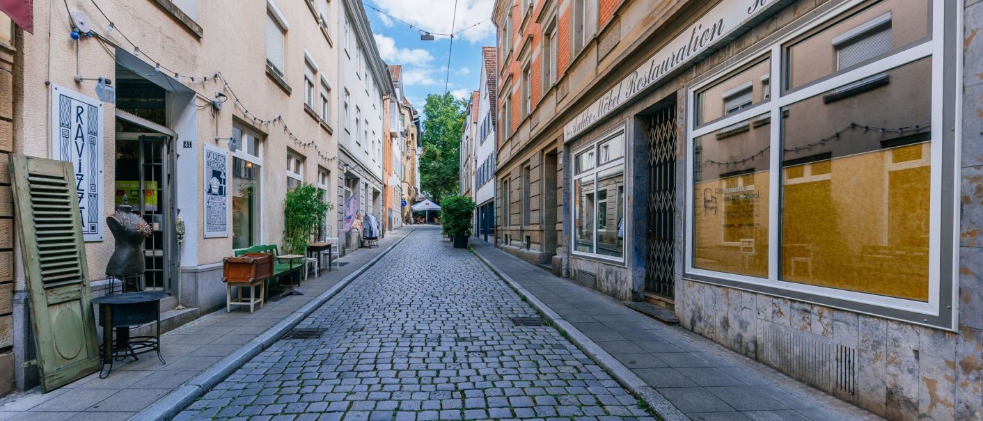 Schmale gepflasterte Straße im Bohnenviertel mit Geschäften und Dekorationen. Links und rechts sind Schaufenster und Pflanzen zu sehen., © Thomas Niedermüller Schmale gepflasterte Straße im Bohnenviertel mit Geschäften und Dekorationen. Links und rechts sind Schaufenster und Pflanzen zu sehen., © Thomas Niedermüller