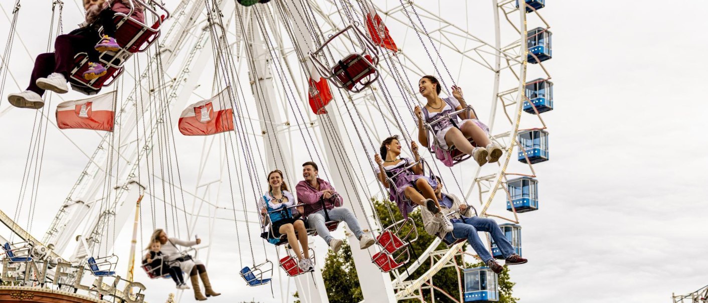 People in traditional costumes ride on a chain carousel in front of a Ferris wheel. Flags wave in the background. Folk festival atmosphere in Stuttgart., &copy; Stuttgart-Marketing GmbH, Sarah Schmid