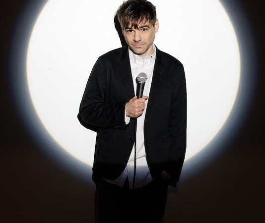 A person stands in the spotlight holding a microphone. The background is dark, the person is wearing a suit and a white shirt., © Rosenau Kultur e.V. A person stands in the spotlight holding a microphone. The background is dark, the person is wearing a suit and a white shirt., © Rosenau Kultur e.V.