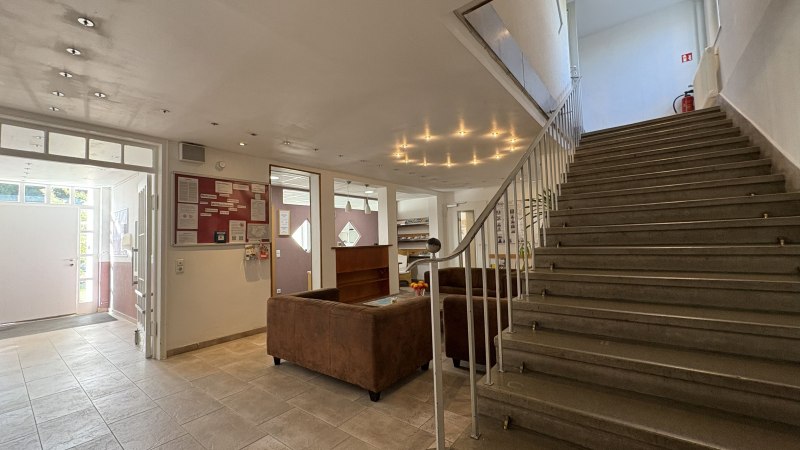 Heller Eingangsbereich mit Treppe, braunen Sofas und Infotafel. Moderne Beleuchtung und Fliesenboden schaffen eine einladende Atmosphäre., © TOMAS Heller Eingangsbereich mit Treppe, braunen Sofas und Infotafel. Moderne Beleuchtung und Fliesenboden schaffen eine einladende Atmosphäre., © TOMAS