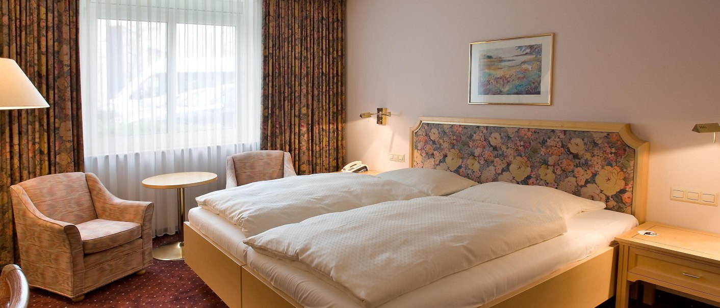 Gemütliches Hotelzimmer mit Doppelbett, floralem Kopfteil, zwei Sesseln, kleinem Tisch und Fenster mit Vorhängen., © TOMAS Gemütliches Hotelzimmer mit Doppelbett, floralem Kopfteil, zwei Sesseln, kleinem Tisch und Fenster mit Vorhängen., © TOMAS
