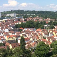 Luftaufnahme einer Stadt mit roten Dächern, einem Fluss und grünen Hügeln im Hintergrund. Der Himmel ist blau mit einigen Wolken., © Aussicht auf den Neckar und die Weinberge