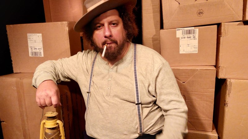 Ein Mann mit Hut und Zigarette sitzt vor gestapelten Kartons und hält eine gelbe Laterne in der Hand., © Theater am Olgaeck Ein Mann mit Hut und Zigarette sitzt vor gestapelten Kartons und hält eine gelbe Laterne in der Hand., © Theater am Olgaeck