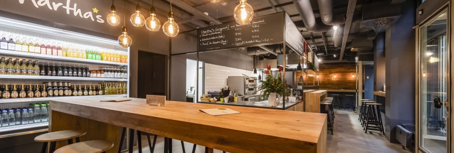 Modernes Café mit Holztischen, Getränkekühlschrank und hängenden Glühbirnen. An der Wand steht "Martha's". Gemütliche Atmosphäre mit Barbereich., © Martha's, Stuttgart