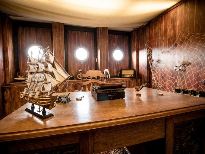 Ein h&ouml;lzerner Schreibtisch mit Modellschiff und Schreibmaschine in einem maritimen Raum. An der Wand h&auml;ngt ein Fischernetz., &copy; ExitGames Stuttgart