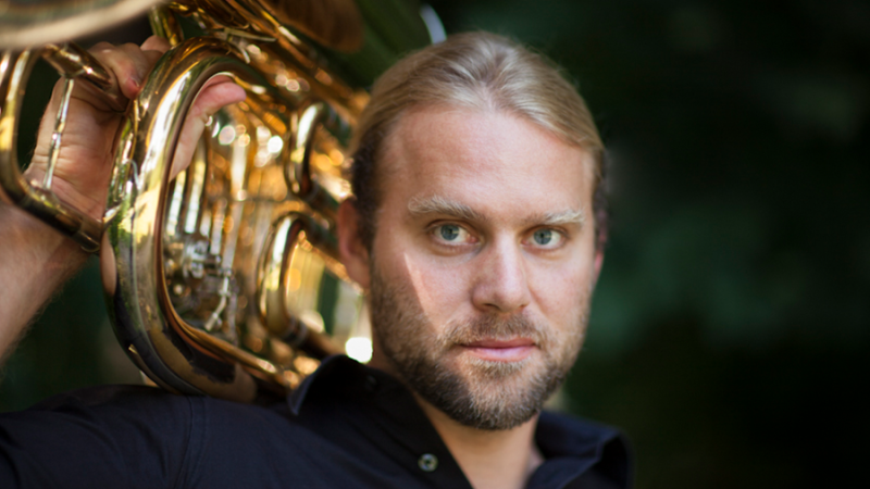 Ein Mann mit blonden Haaren trägt eine Tuba auf der Schulter und blickt ernst in die Kamera. Der Hintergrund ist unscharf und dunkel., © Philippe Gerlach Ein Mann mit blonden Haaren trägt eine Tuba auf der Schulter und blickt ernst in die Kamera. Der Hintergrund ist unscharf und dunkel., © Philippe Gerlach