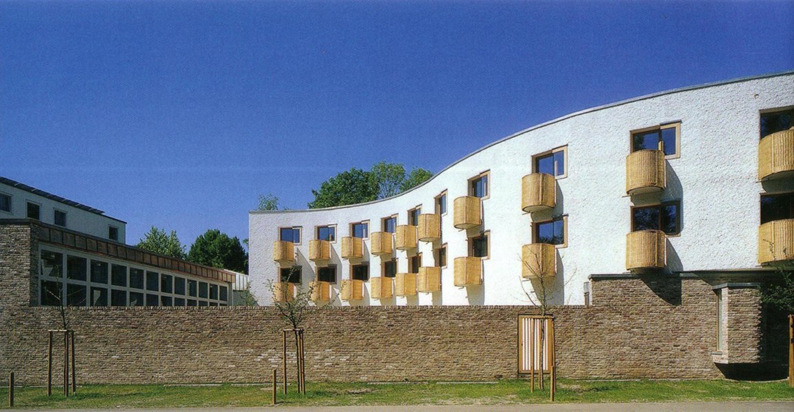 Modernes Gebäude mit wellenförmiger Fassade und runden Balkonen, umgeben von einer Steinmauer, vor blauem Himmel., © TOMAS Modernes Gebäude mit wellenförmiger Fassade und runden Balkonen, umgeben von einer Steinmauer, vor blauem Himmel., © TOMAS