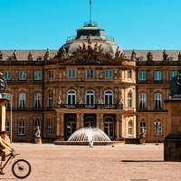 The New Palace in Stuttgart in sunny weather. People walking and a cyclist riding in the foreground. Two statues flank the entrance., &copy; SMG Stuttgart Marketing GmbH - Sarah Schmid