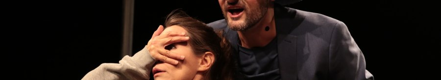 A man in a hat and glasses speaks on stage while covering the eyes of a woman. Both are wearing microphones., © Theater tri-bühne, Stefan Kirchknopf