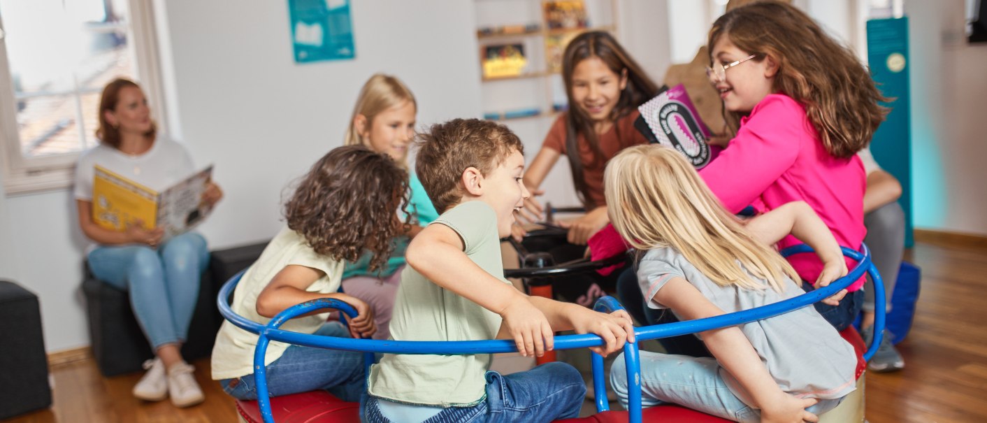 Kinder spielen fr&ouml;hlich auf einem Karussell in einem Raum mit Holzboden. Im Hintergrund sitzt eine Frau mit einem Buch., &copy; FTGRF.de
