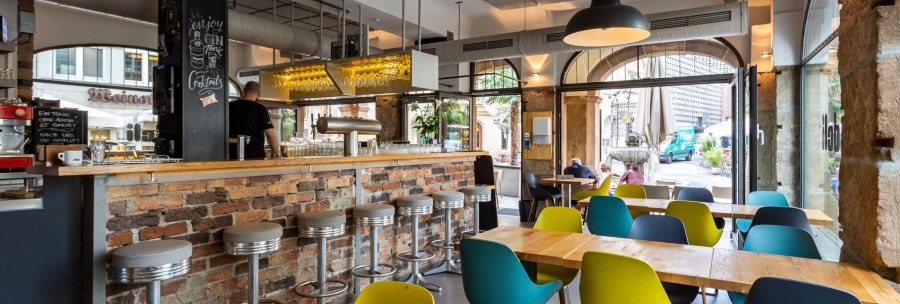 Modernes Café mit Bar aus Backstein, bunten Stühlen und großen Fenstern. Ein Mann steht hinter der Bar, draußen sind Passanten zu sehen., © deli, Stuttgart
