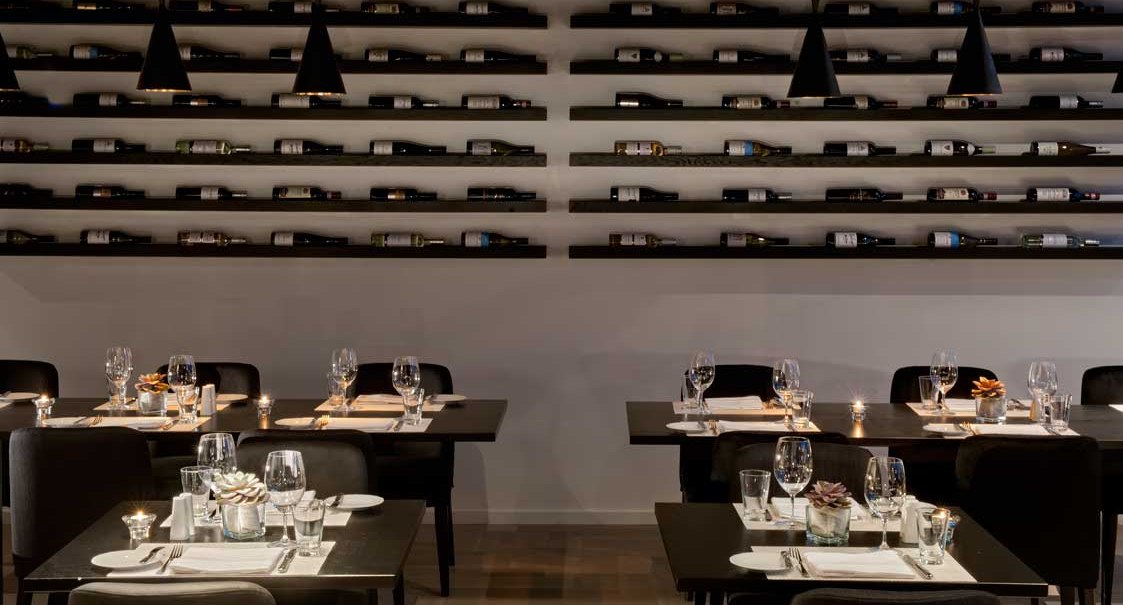 Elegant restaurant with dark tables, set with glasses and cutlery. Wine bottles are arranged decoratively on shelves along the wall., © Wyndham Stuttgart Airport Messe Elegant restaurant with dark tables, set with glasses and cutlery. Wine bottles are arranged decoratively on shelves along the wall., © Wyndham Stuttgart Airport Messe