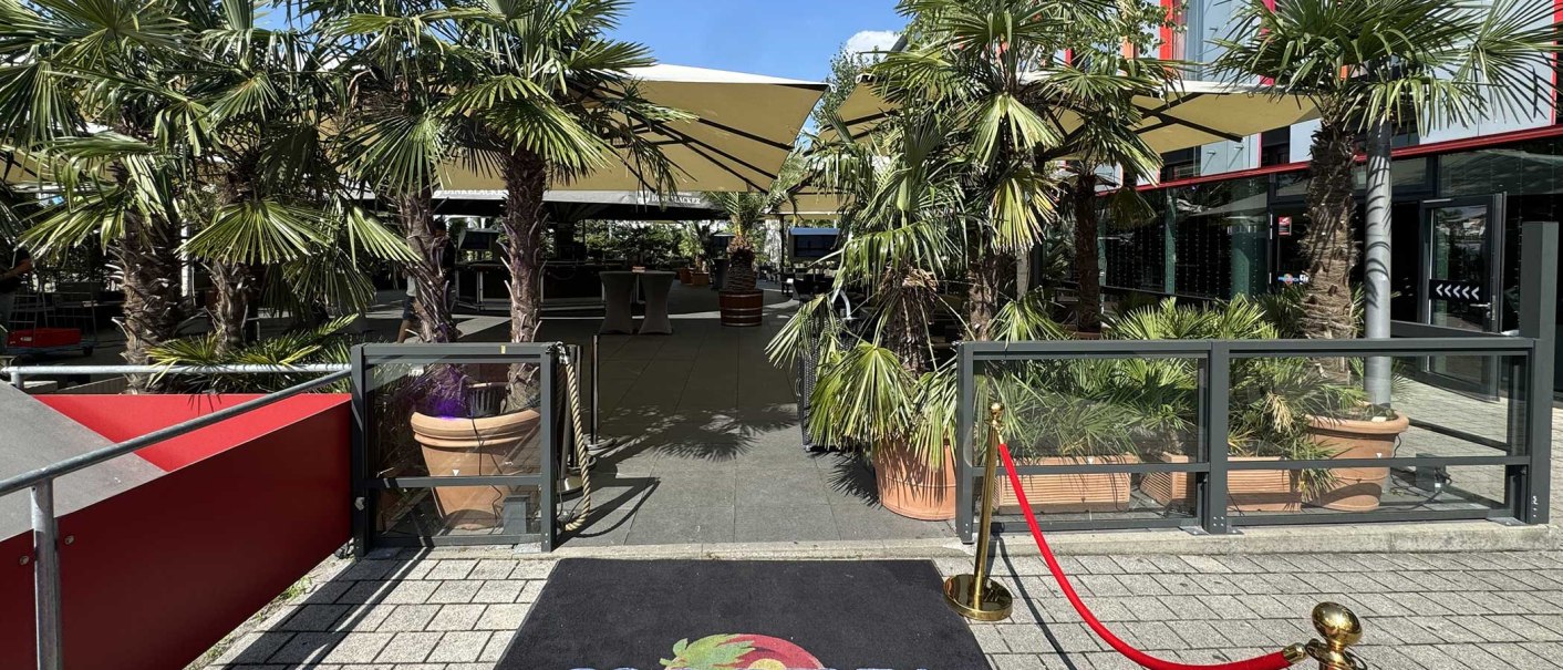 Eingang zu einem Außenbereich mit Palmen, Sonnenschirmen und einem roten Teppich mit 'Palm Beach' Logo. Der Himmel ist blau., © Palm Beach Stuttgart GmbH