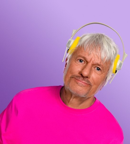 A man with gray hair wears yellow headphones and a pink T-shirt against a purple background., &copy; Renitenztheater Stuttgart e.V.