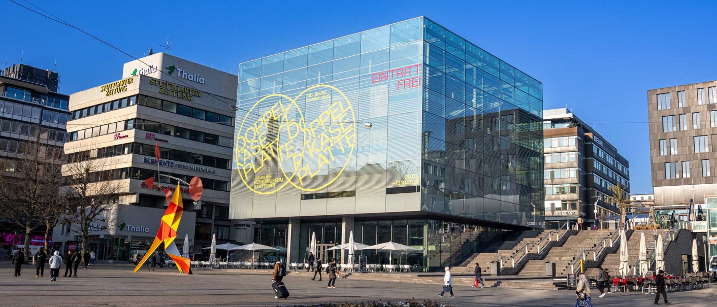 The Stuttgart Art Museum with its striking glass cube on Schlossplatz. A colorful sculpture stands in the foreground, surrounded by modern buildings., &copy; Stuttgart-Marketing GmbH, Sarah Schmid